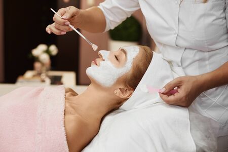 Young beautiful woman during spa, while hands of female cosmetologist applying peel mask to her face...