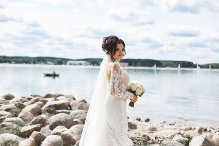 Young bride with a wedding hairstyle in a white lace dress stands on the coast of the lake. Young woman in a wedding dress with a bouquet of flowers posing near the lake in spring day. Wedding fashionの写真素材