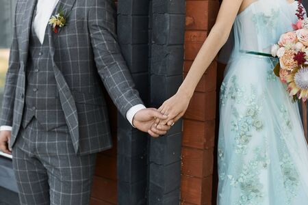 Cropped portrait of a handsome young man in checkered suit holding the hand of young woman in beige dress. Modish groom and stylish bride holding hands. Closeup photo of two young lovers holding handsの写真素材