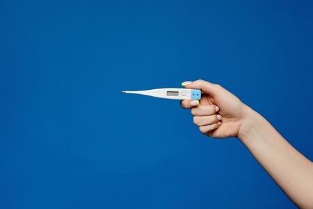Electronic medical thermometer in a neat female hand with manicure isolated on a blue background...