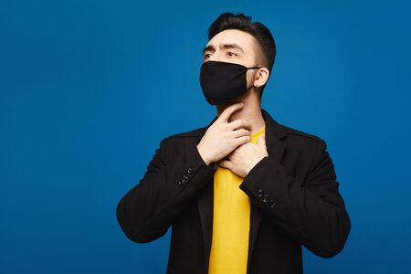 Young man in black protective mask with throat sore, isolated. Handsome men holds hands on his neck because of throat pain. Inflamed throat. Portrait of sick man in medical mask touching painful neck.の写真素材
