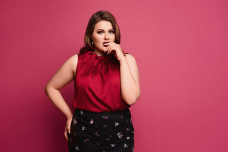 Pensive plus-size model girl with bright makeup and with stylish hairstyle in a red blouse and skirt posing at the pink background, isolatedの写真素材