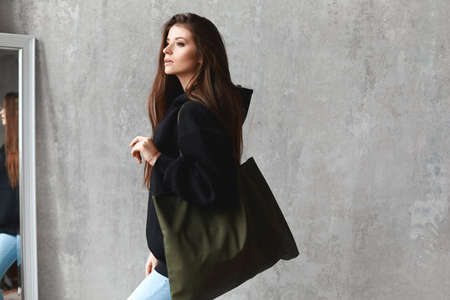 A young woman in a black sweatshirt and blue jeans with a big bag on her shoulder posing indoorsの写真素材