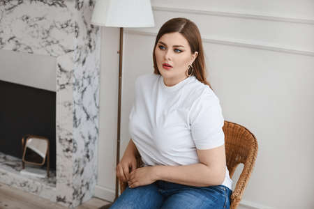 Plus size model girl in jeans and blank white t-shirt sits in the armchair in the living room. XXL...