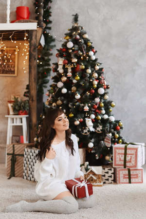 Young woman with slim body in knitted dress keeps Christmas gift and sitting near the Christmas tree.の写真素材
