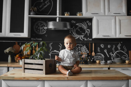 Toddler boy in a white shirt and shorts looking aside while sitting on the kitchen tableの写真素材