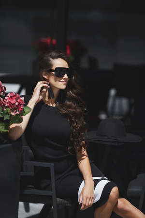 Outdoor summer portrait of a model woman wearing trendy sunglasses and fashionable dress sitting at the cafe table in the street of a European cityの写真素材