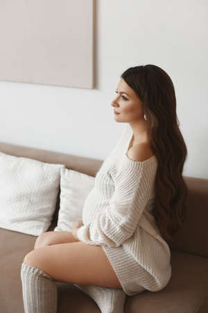 Beautiful young pregnant woman with dark hair wearing white knitted dress posing on the sofaの写真素材