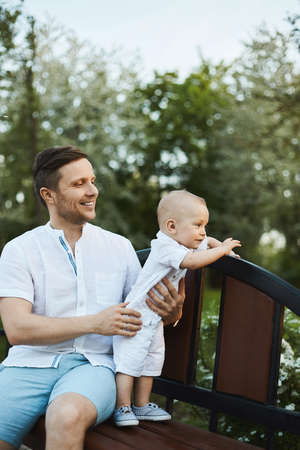 A handsome young man and adorable toddler boy, young dad and his little son, posing on the bench at a city park on a warm summer dayの写真素材
