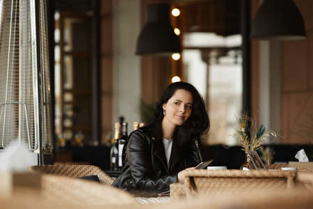 Portrait of a young female model with dark hair wearing leather jacket sitting at the table in a cafeの写真素材