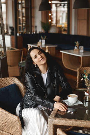 Pensive European female student sitting at the table in a cafe, drinking coffee and using her...