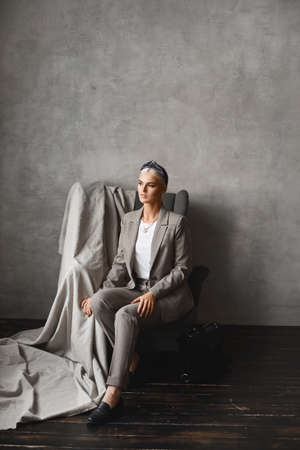 A young Caucasian woman with blond hair looking aside while sitting in the armchair. Gorgeous young businesswoman in stylish suit posing in an empty interiorの写真素材