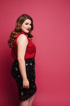 Young plus-size model girl in red satin blouse and skirt posing over pink background. Happy chubby...
