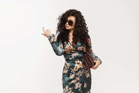 A young brunette woman with curly hair in trendy dress pointing her finger up and posing over white background. Model girl in fashionable clothes isolated on white backgroundの写真素材