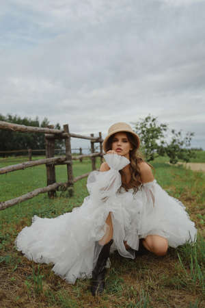 Beautiful model girl in trendy vintage dress and straw hat at the countryside. Young bride in modish dress posing outdoors at the rural. Concept of wedding fashionの写真素材