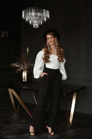 Fashionable young woman in white blouse and trendy hat posing in dark luxurious interiorの写真素材