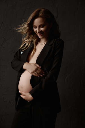 A gorgeous pregnant young woman with fluttering blond hair wearing an unbuttoned blazer embracing her belly and posing beside the black wall. Naked belly. Lifestyle, pregnancy, healthの写真素材