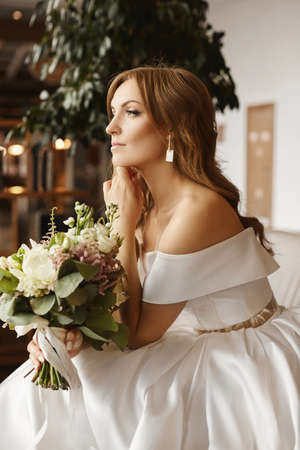 Pensive stunning bride sitting with a bridal bouquet in waiting of groom. Attractive young woman in fashionable white dress with a bouquet of fresh flowersの写真素材