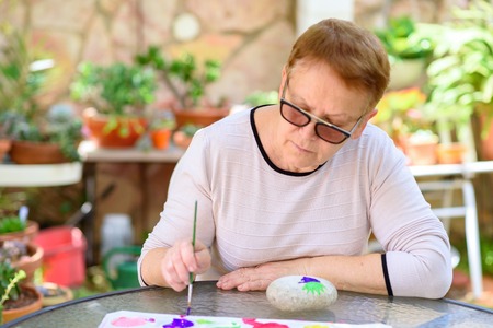 Happy authentic senior woman sitting outdoors at table painting pictures on stone.の写真素材