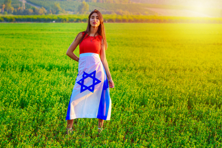 Young woman patriot jewish proud girl standing wrapped flag of Israel on amazing landscape in beautiful summer or spring day.Beautiful Brunette happy teen walk outdoor wearing gorgeous red dress .の写真素材