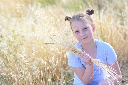 Beautiful girl holding spikes of wheat and ears of oats. Cute child sitting on the gold autumn field ready for harvest.の写真素材