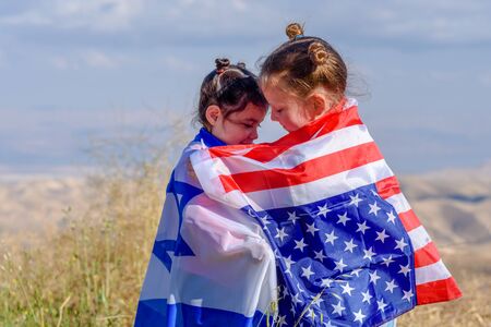 Two cute girls with American and Israel flags. Little children holding Israeli and USA flags hugging on meadow with beautiful landscape in background.の写真素材