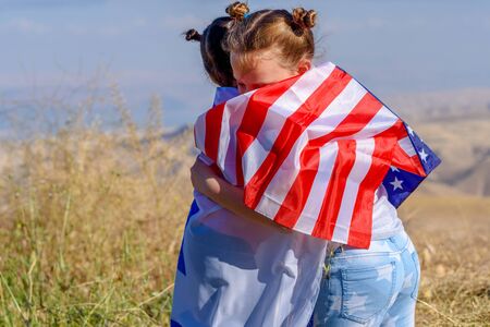 Two cute girls with American and Israel flags. Little children holding Israeli and USA flags hugging on meadow with beautiful landscape in background.の写真素材