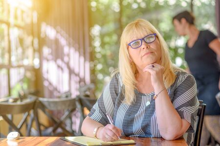 Portrait of beautiful plus size middle age woman working and making notes to her diary on sunny terrace of cafe. Elegant middle aged lady posing with modern glasses.