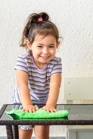 Cute laughing curly toddler girl washing glass table with green towel in home terrace. Sunny day.の写真素材