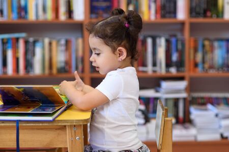 Little Girl Indoors In Front Of Books. Cute Young Toddler Sitting On A Chair Near Table and Reading...