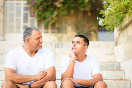 Teenager son and senior father sitting on stairs outdoors at home, talking.の写真素材