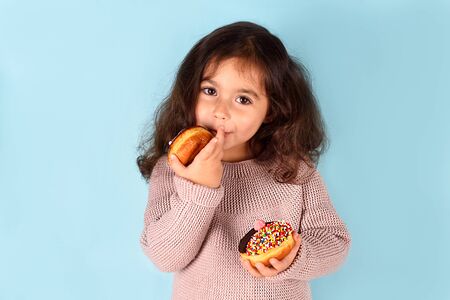 Little happy cute girl with curly hair eating donut licks her fingers, on which sugar is stuck.
