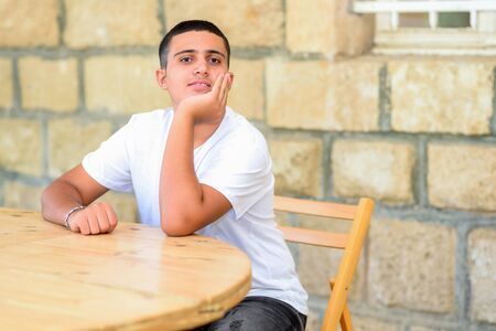Black hair teenage boy sitting in high school yard at the table. Male student in college outdoor classroom.の写真素材