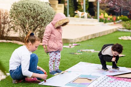 Kids play outdoors. Children paints a paper house template with colorful watercolors on the grass in the garden.の写真素材