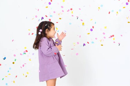 Portrait of a beautiful little girl wearing dress standing under confetti rain and celebrating over white background.の写真素材