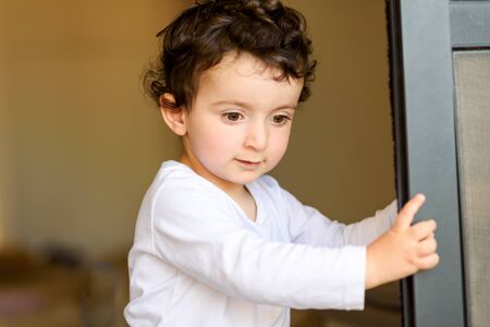 Funny boy with curly hair behind door at home. Photo of adorable young happy kid looking down....