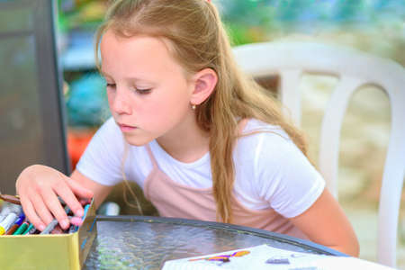 Little blonde girl painting at table in summer garden. Cute freckles child drawing outdoor.Creative kid doing homework at backyard, choosing a pencil.Having fun on a summer vacation , holiday concept.の写真素材