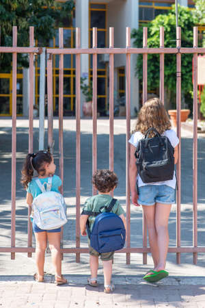 A Young Sisters And Brother Wait Outisde A Closed School Gate. Diverse school children going to school.の写真素材
