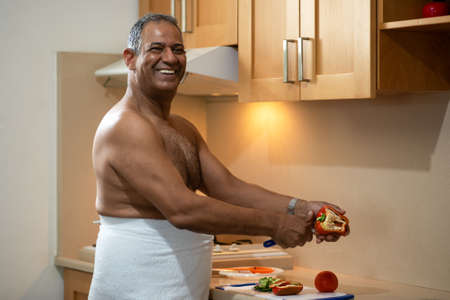 Perfect fit dark-skinned smiling happy senior man in the white towel looking at camera while standing in the kitchen and cutting salad.の写真素材