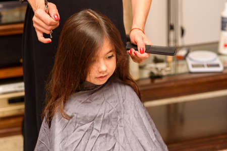 Portrait of a young girl getting her hair cut in home. Moms hand with hairdressing scissors. Little child is getting a haircut in home during the lockdown.の写真素材