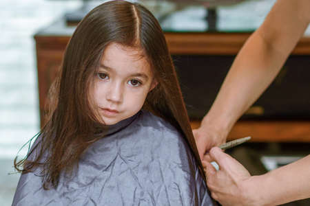 Portrait of a young girl getting her hair cut in beauty hair salon. Womans hand with hairdressing scissors. Little child is getting a haircut in home during the lockdown.の写真素材