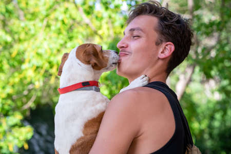 Portrait of a young man holding his jack Russell terrier dog in his arms. Cute dog kisses the face of owner in black tank top.の写真素材