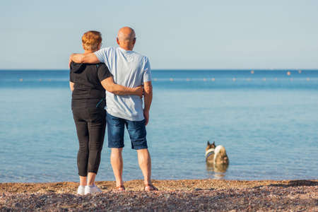 Happy senior couple hugging on summer beach. Back view of sustainable eco-friendly regenerative travel old tourists hiker walking on a beach enjoying healthy environment renewable resources.の写真素材