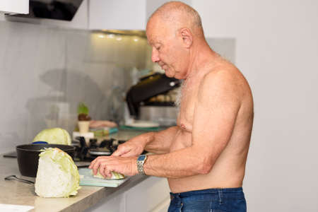 Senior man at home is cooking. Extreme heat summer, shirtless mature man inside home in his kitchen trying to cool off and prepares vegetables.の写真素材