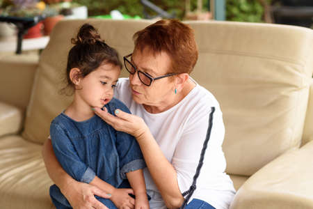 Little girl talking to her grandma sitting on the couch. Portrait of loving grandmother and granddaughter together at backyard. Little child is sitting on grand moms lap.の写真素材