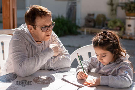 Beautiful girl doing homework, writing and reading with the help of her grandmother on an outdoor pergola. Close up portrait of senior teacher supervising little kid doing homework back yard.の写真素材