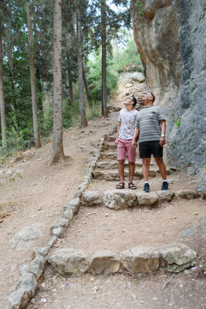 Happy teen son and senior father walking in woods. Adult dad and young child looking up to the trees...