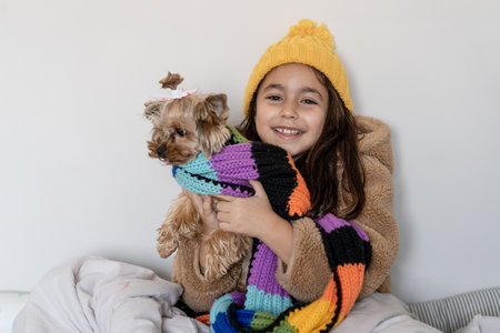 Child in warm faux fur coat, knitted hat and scarf sitting with funny small dog in a bed in home in cold winter time.Beautiful little girl warms in blanket and looks the camera.Selective focus on kid.の写真素材