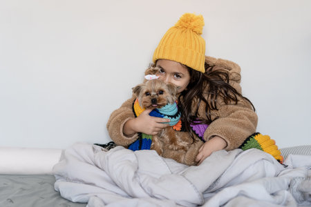 Child dressed in beige warm faux fur coat and knitted hat and scarf sitting with funny small dog in...