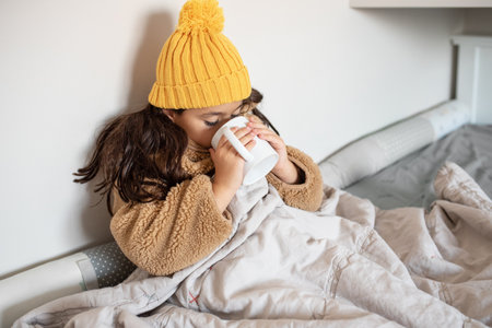 Beautiful little girl warms in blanket in cold winter time. Child in warm clothes sitting in a bed drinking hot tea at home. Cold home. No heating.の写真素材